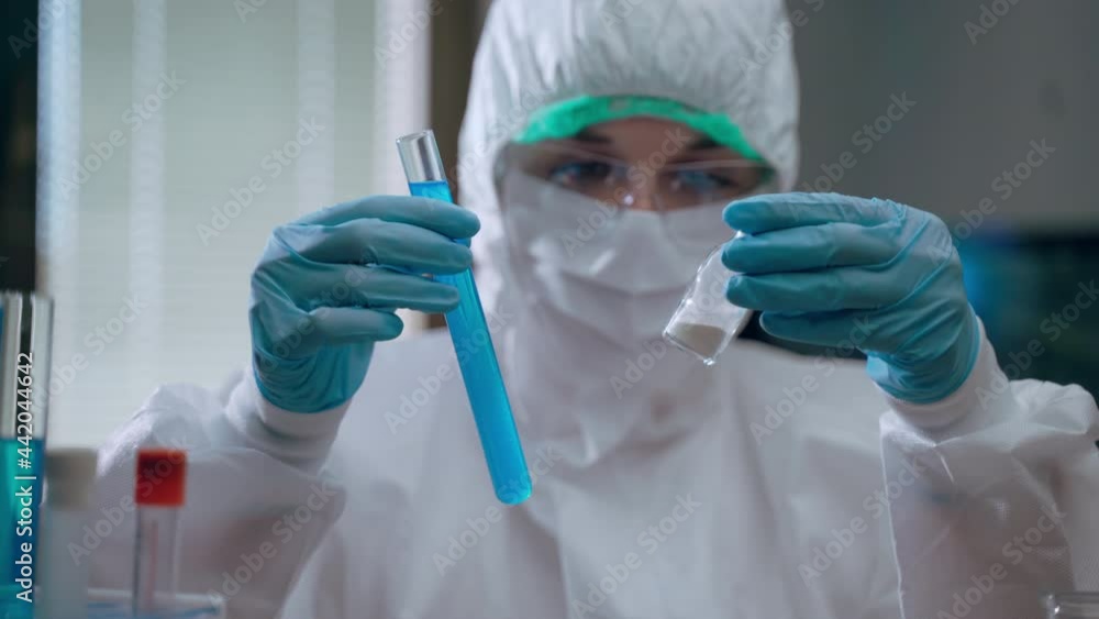 Close-up of female virologist worker in protective coverall holding sterile test tube with blue reagent in one hand and vial with powder in another one, laboratory researcher working with hazard Close-up of female virologist worker in protective coverall holding sterile test tube with blue reagent in one hand and vial with powder in another one, laboratory researcher working with hazard