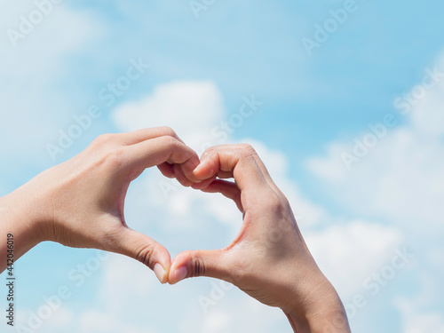Heart shape hand on blue sky and fluffy cloud background, concept of love, relationship and romantic. Female hand making finger love sign with copy space. © tete_escape