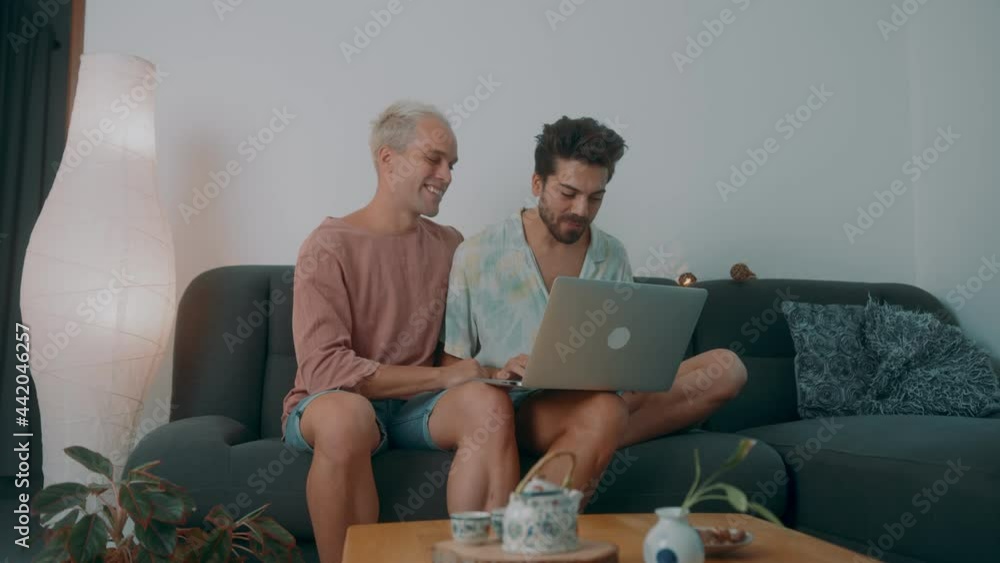 A European gay couple spending time at home. They lie on the couch and use their laptop A European gay couple spending time at home. They lie on the couch and use their laptop
