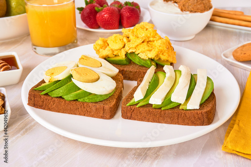 3 toasted breads with avocado  hard boiled egg and scrambled eggs on a white plate  accompanied by Greek yogurt and fresh fruit on a marble surface and white background. close up