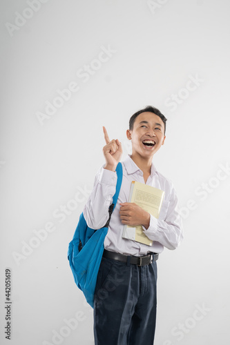 a boy in junior high school uniform smiling looking up with finger pointing when carrying a book and a backpack with copyspace on an isolated background