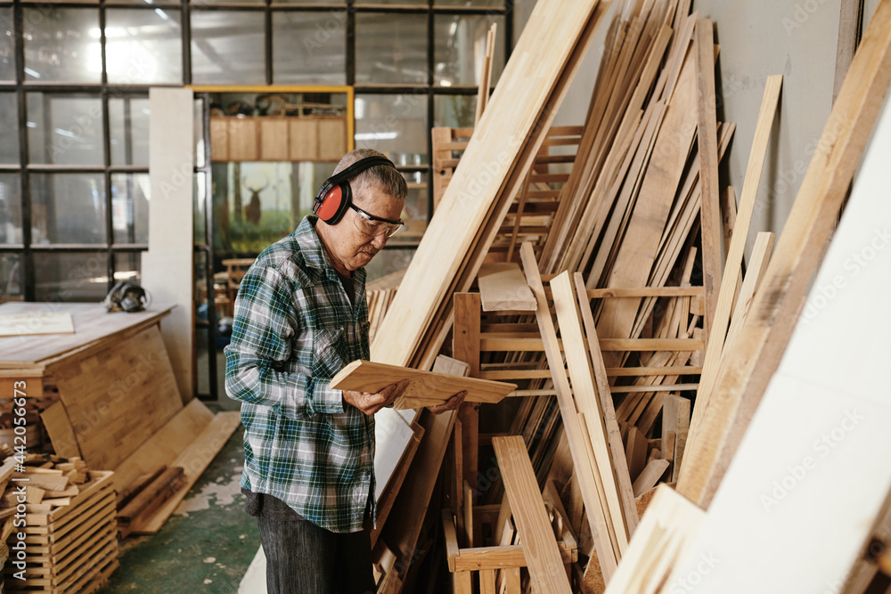 Serious experienced elderly carpenter choosing wooden piece for carving or making furniture item Serious experienced elderly carpenter choosing wooden piece for carving or making furniture item