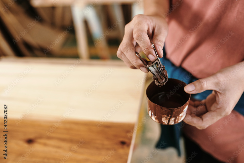 Fototapeta premium Close-up imege of female carpenter soaking binder clip with piece of cloth in jar with oil to cover wooden surface
