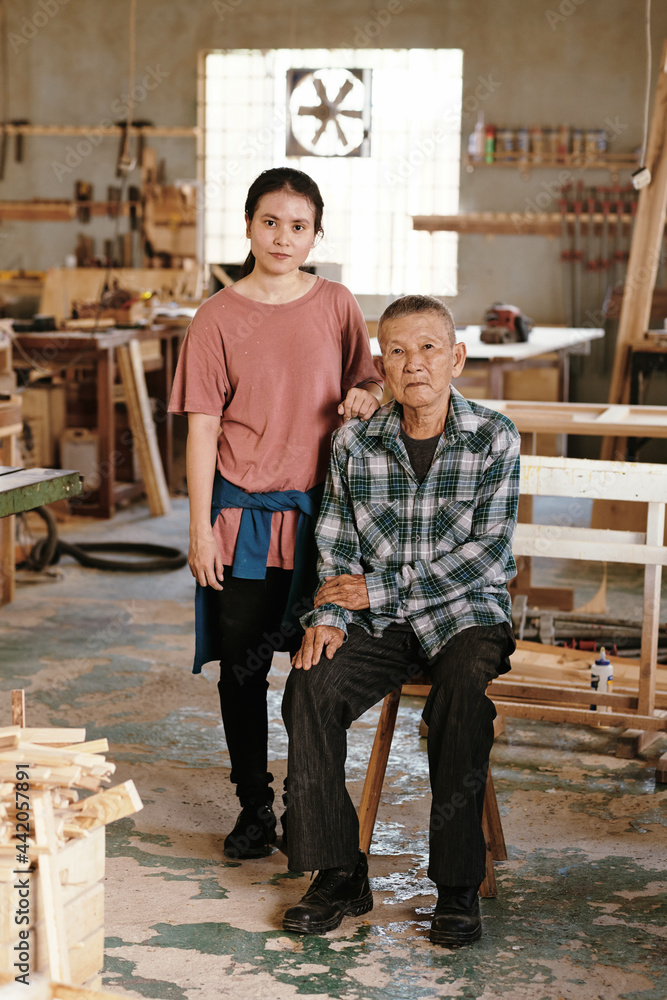 Portrait of serious senior carpenter and his granddaughter standing in family carpentry workshop