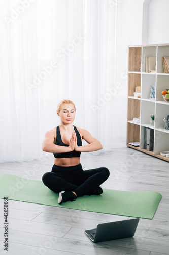 Home yoga practice. Inspired woman. Online training. Healthy body. Peaceful mind. Meditating woman black sportswear sitting lotus pose namaste hands looking laptop in light room interior.