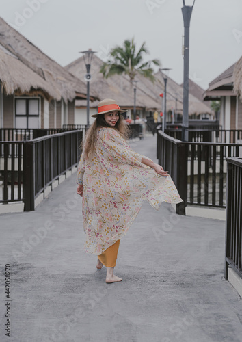 Back view of a girl with beautiful long hair wearing an orange dress and a straw hat walking gracefully in a summer luxury resort. 