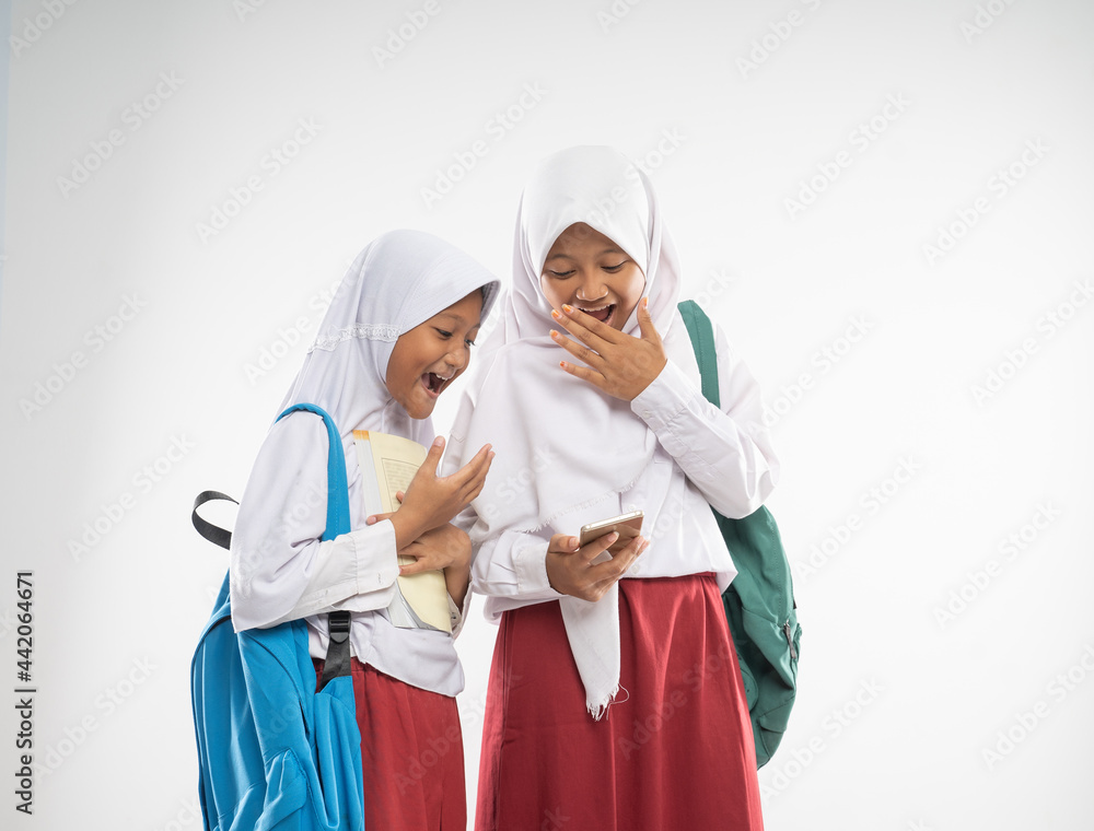 two veiled girls wearing elementary school uniforms using a mobile phone together with shocked expression when carrying backpack an isolated background two veiled girls wearing elementary school uniforms using a mobile phone together with shocked expression when carrying backpack an isolated background