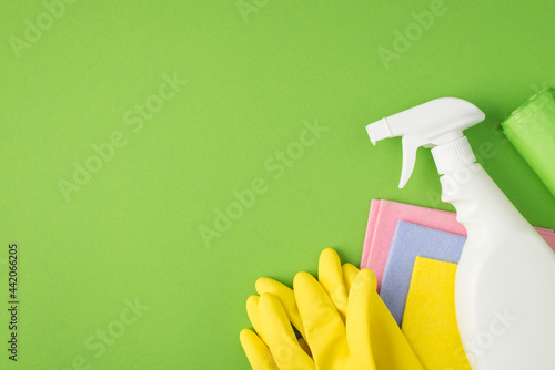 Top view photo of white spray detergent bottle without label multicolor viscose rags garbage bags and yellow rubber gloves on isolated green background with copyspace