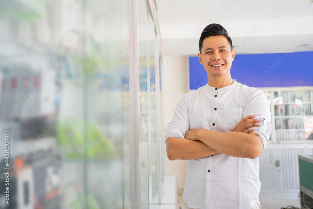 handsome asian man smiling with crossed hands while standing near cell phone accessories display case in cell phone shop handsome asian man smiling with crossed hands while standing near cell phone accessories display case in cell phone shop