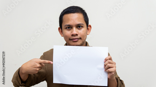 A portrait of young Asian Malay man with casual brown shirt holding and pointing to an empty blank paper on isolated white backgrounds. Empty space for text.