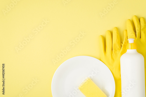 Top view photo of detergent foam bottle without label yellow sponge on dish and rubber gloves on isolated yellow background with blank space