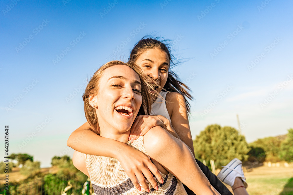 Two girls best friends having fun outdoor in a green nature park looking at camera laughing and joking. Young brunette woman jumping on back of her blonde girlfriend surprising her. Happy students Two girls best friends having fun outdoor in a green nature park looking at camera laughing and joking. Young brunette woman jumping on back of her blonde girlfriend surprising her. Happy students
