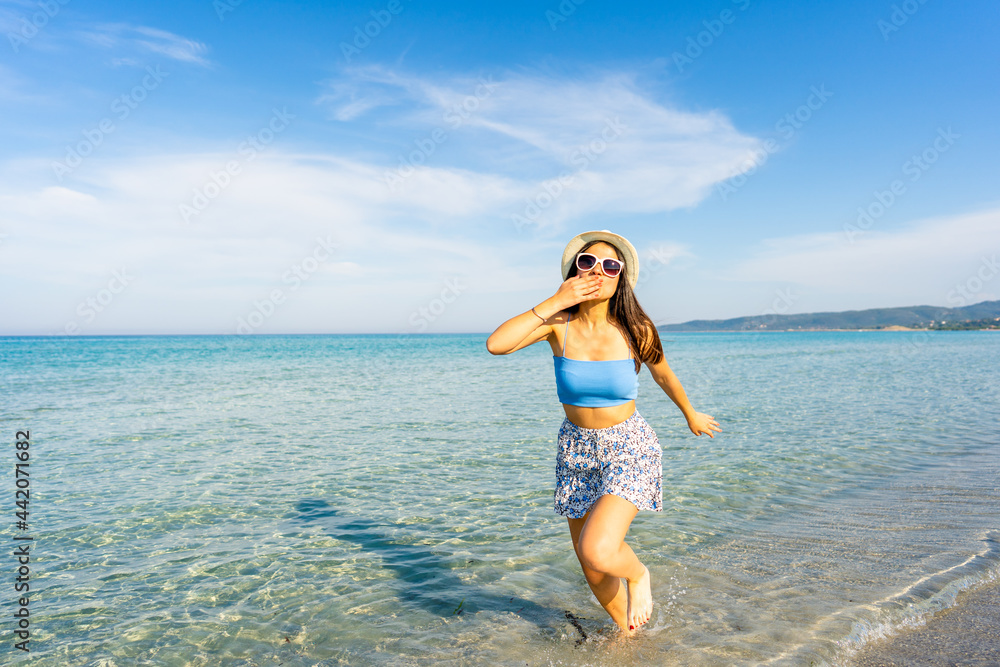 Young beautiful woman making kiss gesture with hand on mouth looking at camera walking in the seawater wearing white hat and sunglasses. Spend your time in travel for a happy better life experience Young beautiful woman making kiss gesture with hand on mouth looking at camera walking in the seawater wearing white hat and sunglasses. Spend your time in travel for a happy better life experience
