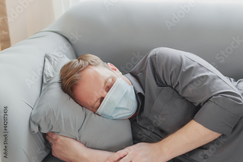 Young attractive Caucasian men relaxing wearing protective face mask having a nap on couch in the afternoon. Happy employee man resting with eyes closed on the sofa during state quarantine