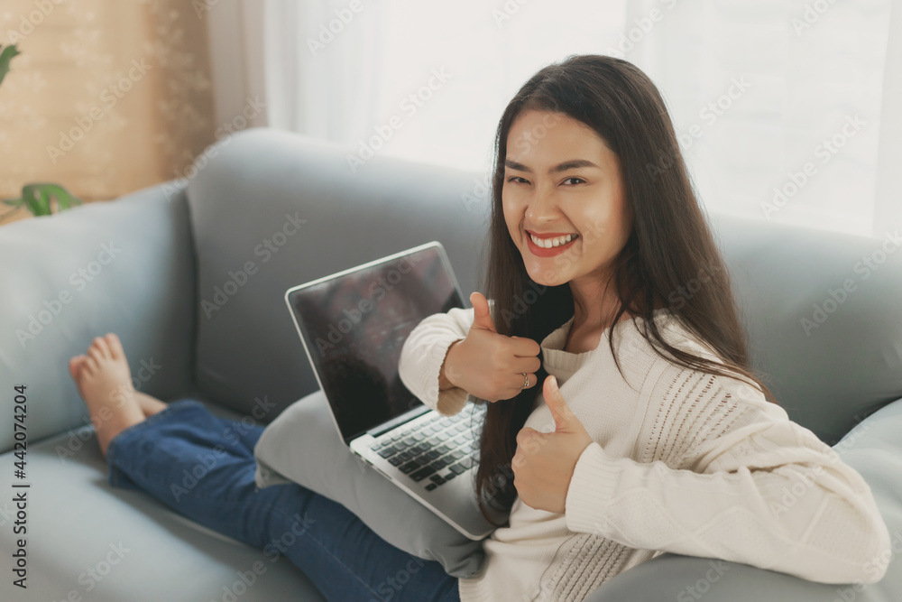 Young happy Asian wearing casual clothes using laptop computer relax sitting on couch sofa at home. Asian employee with Success expression showing thumps up looking at camera with smiley face Young happy Asian wearing casual clothes using laptop computer relax sitting on couch sofa at home. Asian employee with Success expression showing thumps up looking at camera with smiley face