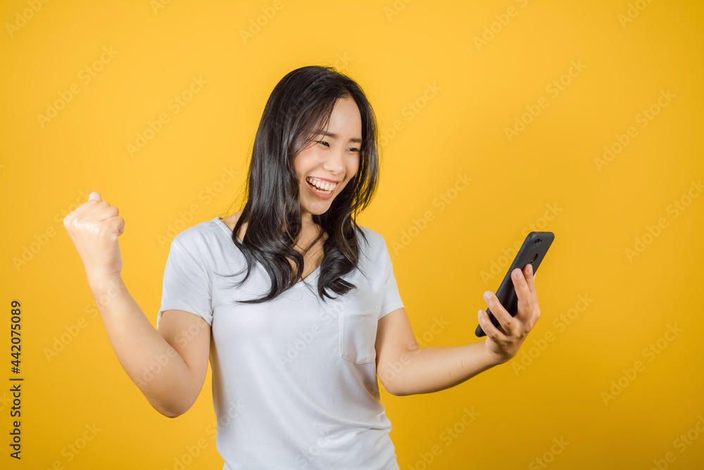 Portrait of happy Asian woman wears white t-shirt raise hands glad excited cheerful and holding mobile phone or smartphone isolated on bright yellow background Portrait of happy Asian woman wears white t-shirt raise hands glad excited cheerful and holding mobile phone or smartphone isolated on bright yellow background