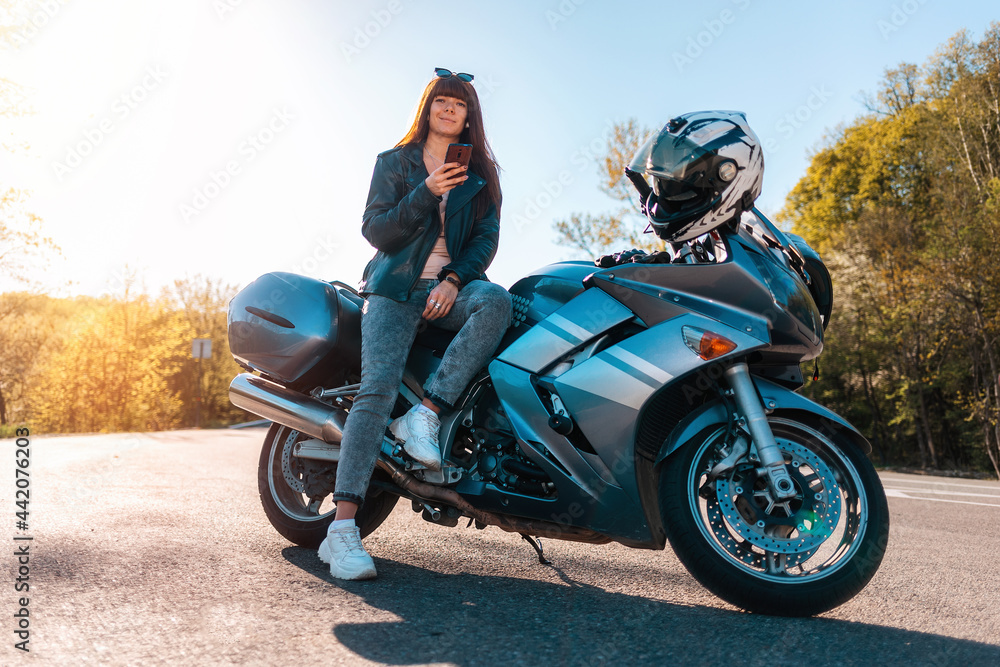 Obraz premium Motorcycle local travel. Young smiling woman in a leather jacket and sunglasses sitting on a motorbike and using a smartphone. World Motorcyclist Day concept