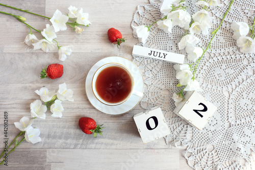 Calendar for July 2 : cubes with the numbers 0 and 2, the name of the month of July in English, a cup of tea on a gray openwork napkin, scattered flowers of bluebells and strawberries, top view