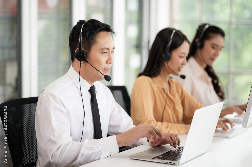 Team of call center staff in Asia wears headphones with a microphone. Smile while serving customers at desks and computers. Service concept and consulting. Communication concept. Team of call center staff in Asia wears headphones with a microphone. Smile while serving customers at desks and computers. Service concept and consulting. Communication concept.