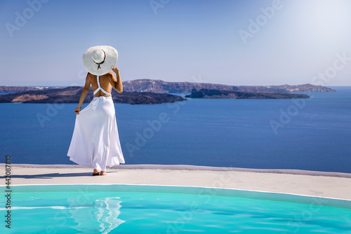 A beautiful woman in a white summer dress stands by the pool and enjoys the breathtaking view over the mediterranean sea