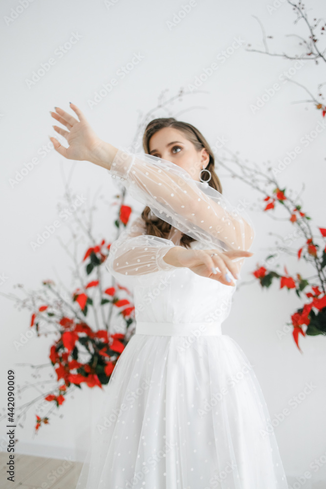 A beautiful young girl with brown hair in a white wedding dress with sleeves. A bride with her hair down in a photo studio. the decor is made of red flowers. the concept of a festive image. A beautiful young girl with brown hair in a white wedding dress with sleeves. A bride with her hair down in a photo studio. the decor is made of red flowers. the concept of a festive image.