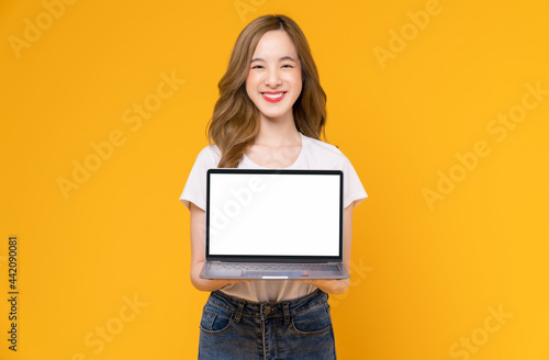 Studio shot of cheerful beautiful Asian woman in white t-shirt and holding laptop mockup of blank screen on yellow background.