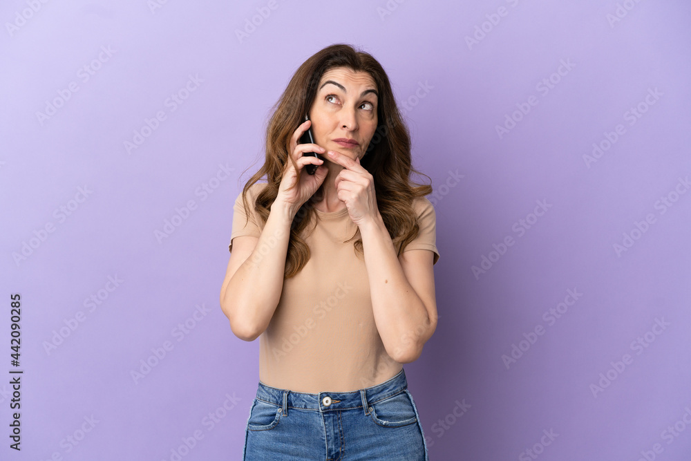 Middle aged caucasian woman using mobile phone isolated on purple background looking up while smiling Middle aged caucasian woman using mobile phone isolated on purple background looking up while smiling