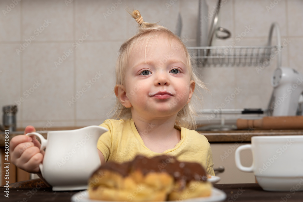 Blonde child is having breakfast in the kitchen. Kid drinking tea. Little girl drinking tea with sweets Blonde child is having breakfast in the kitchen. Kid drinking tea. Little girl drinking tea with sweets