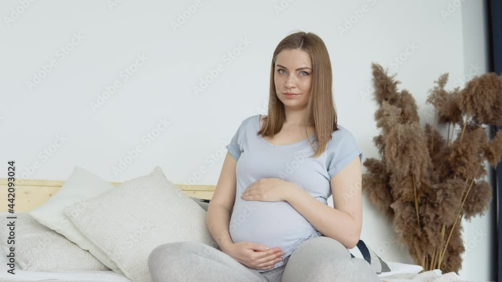 Pregnant woman in home clothes sitting on a double bed. Dried flowers in the interior. The pregnant woman is stroking her belly. Third trimester of pregnancy Pregnant woman in home clothes sitting on a double bed. Dried flowers in the interior. The pregnant woman is stroking her belly. Third trimester of pregnancy