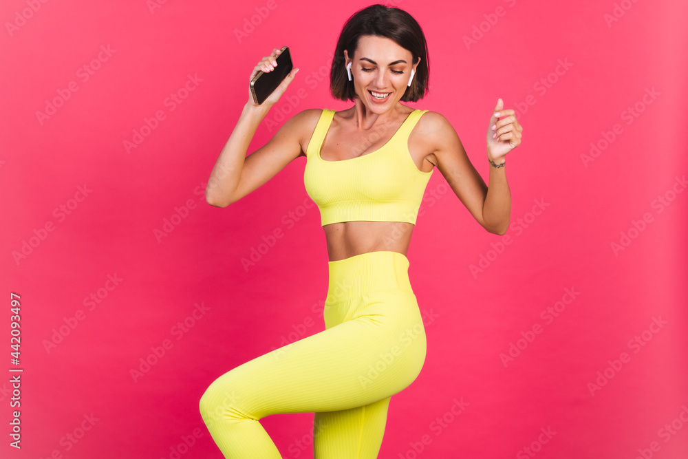 Beautiful fit woman in yellow bright fitting sportswear on pink background happy dancing headphones in ears hold phone listen music excited perfect body Beautiful fit woman in yellow bright fitting sportswear on pink background happy dancing headphones in ears hold phone listen music excited perfect body