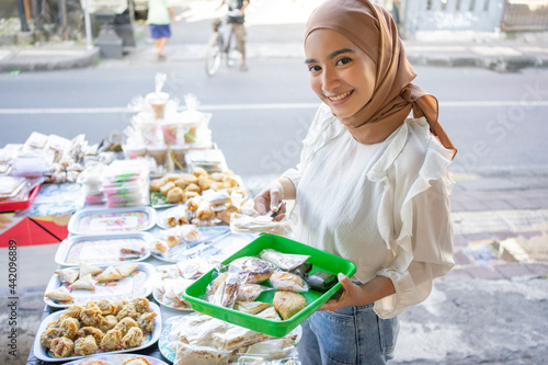 A beautiful girl wearing a headscarf uses tongs and carrying a plastic tray choosing snacks to buy at a roadside stall