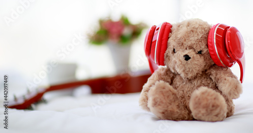 in home bedroom, brown lovely fluffy teddy bear toy creatively wearing red earphone and sit on bed near guitar as lovely doll for enjoy music playing on delight celebration holiday