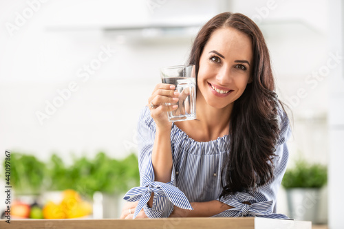 Smiling pretty woman holds a glass of water leaning on kitchen desk
