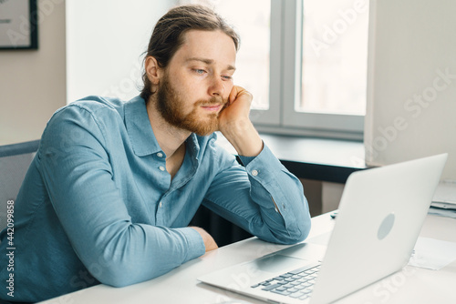 Young unhappy man office worker feeling bored at work, looking at laptop with demotivated face expression while sitting at workplace in office, distracted male worker feeling tired of monotonous job
