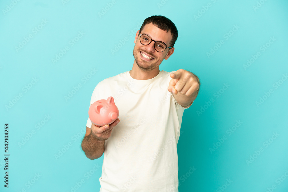 Brazilian man holding a piggybank over isolated blue background pointing front with happy expression Brazilian man holding a piggybank over isolated blue background pointing front with happy expression