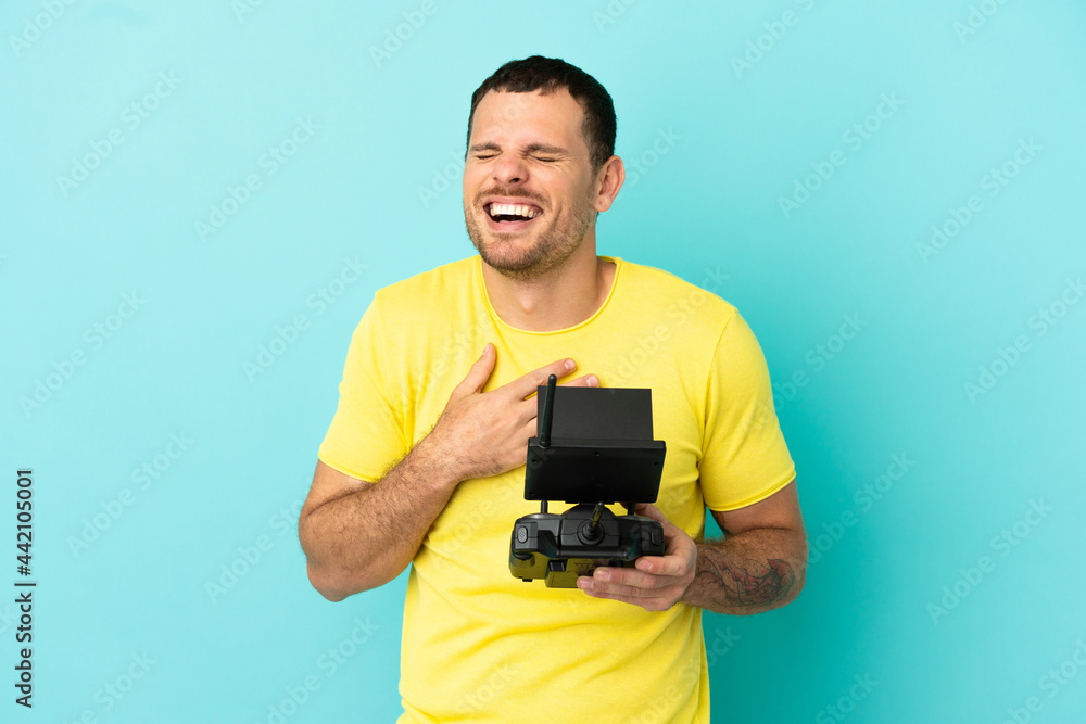 Brazilian man holding a drone remote control over isolated blue background smiling a lot Brazilian man holding a drone remote control over isolated blue background smiling a lot
