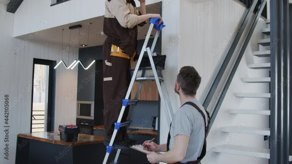 Tilt up slowmo shot of handyman writing on clipboard while electrician in uniform standing on ladder and inspecting smart home device or sensor Tilt up slowmo shot of handyman writing on clipboard while electrician in uniform standing on ladder and inspecting smart home device or sensor