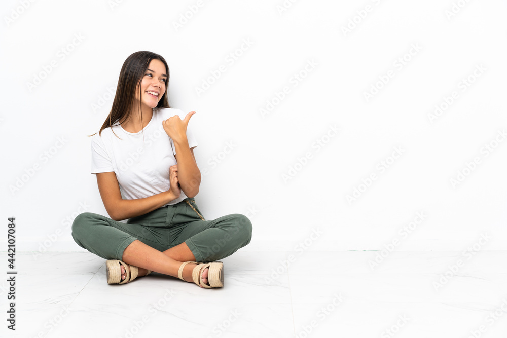 Teenager girl sitting on the floor pointing to the side to present a product Teenager girl sitting on the floor pointing to the side to present a product