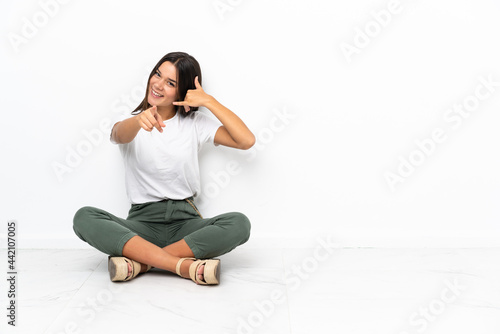 Teenager girl sitting on the floor making phone gesture and pointing front
