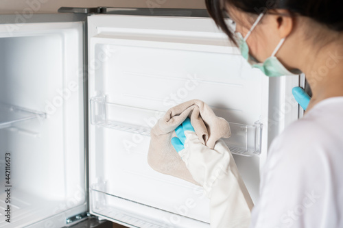 Young Asian woman wearing medical mask and hands in white rubber protective glove cleaning white empty new refrigerator with rag. Cleaning service, housewife, routine housework concept.