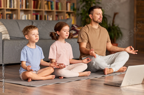 Dad with child little son and daughter doing yoga online  sitting in lotus pose on floor and meditating with closed eyes