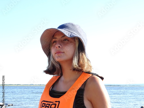 retrato rostro chica adolescente  rubia de pelo corto lacio con pecas ojos castaños en la playa con sombrero pituso azul y chaleco salvavidas naranja de fondo el agua de un lago photo