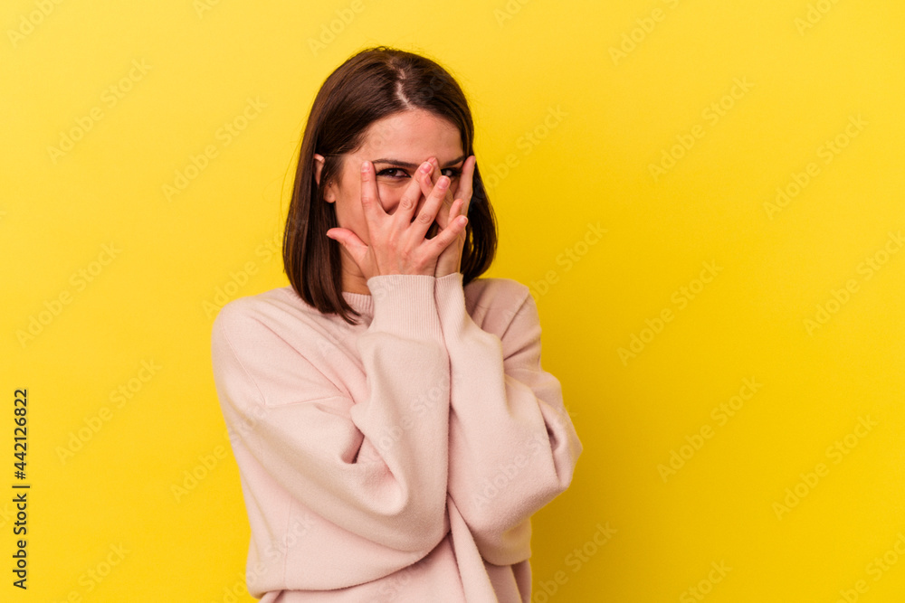 Young caucasian woman isolated on yellow background blink at the camera through fingers, embarrassed covering face. Young caucasian woman isolated on yellow background blink at the camera through fingers, embarrassed covering face.
