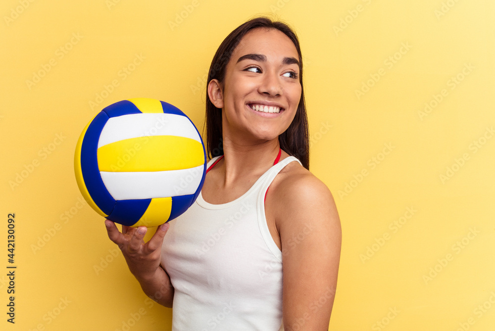 Young mixed race woman playing volleyball on the beach isolated on yellow background looks aside smiling, cheerful and pleasant.