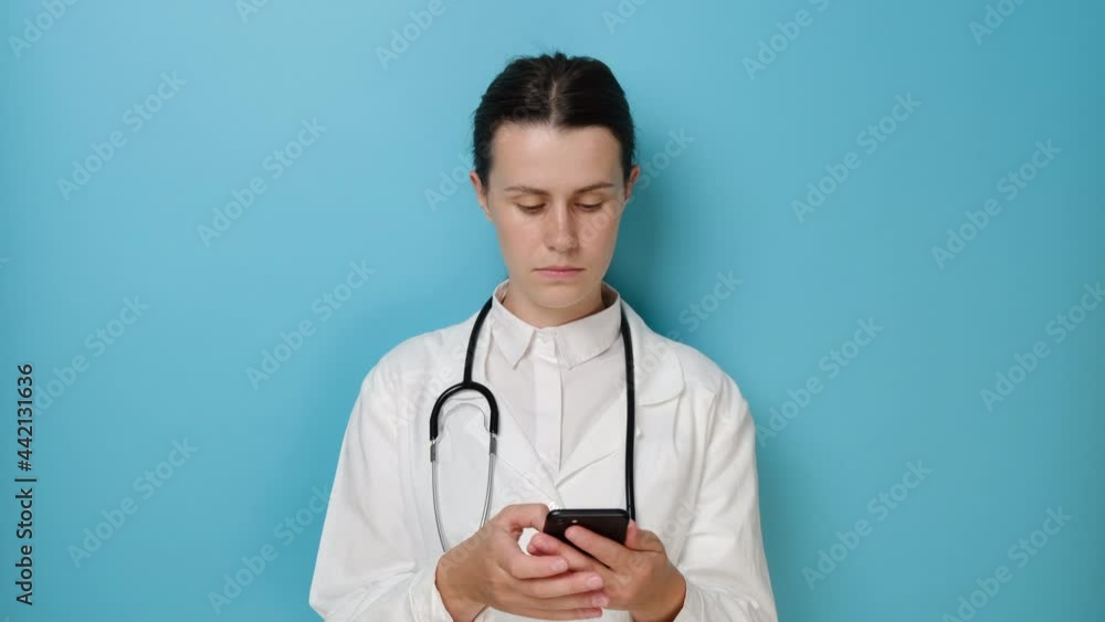 Happy woman doctor in white coat standing isolated on blue studio background hold smartphone, check messages from patient, use mobile app for medical workers easy comfort usage of modern tech concept Happy woman doctor in white coat standing isolated on blue studio background hold smartphone, check messages from patient, use mobile app for medical workers easy comfort usage of modern tech concept