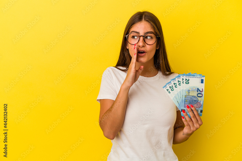 Young caucasian woman holding bills isolated on yellow background is saying a secret hot braking news and looking aside Young caucasian woman holding bills isolated on yellow background is saying a secret hot braking news and looking aside