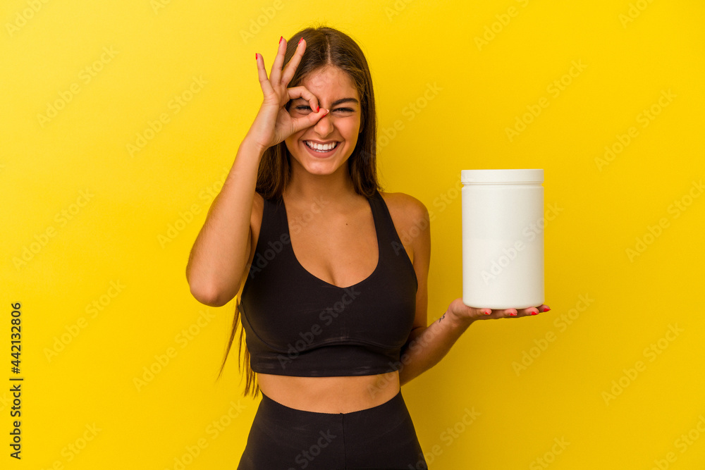Young caucasian woman holding a protein bottle isolated on yellow background excited keeping ok gesture on eye.