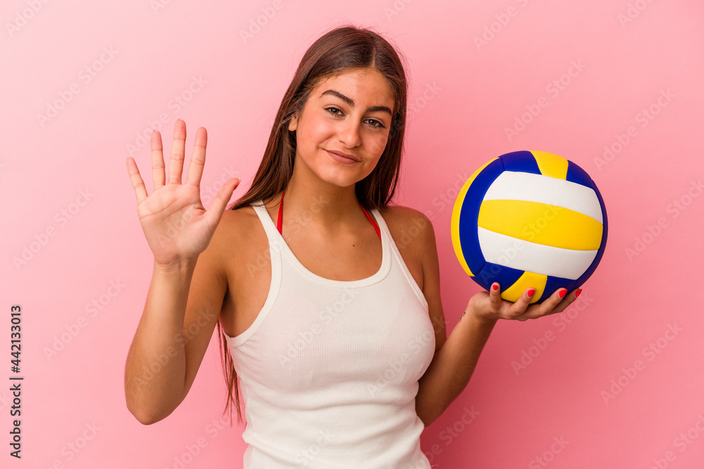 Young caucasian woman holding a volleyball ball isolated on pink background smiling cheerful showing number five with fingers.