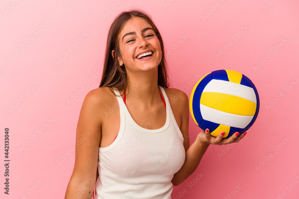 Young caucasian woman holding a volleyball ball isolated on pink background laughing and having fun.