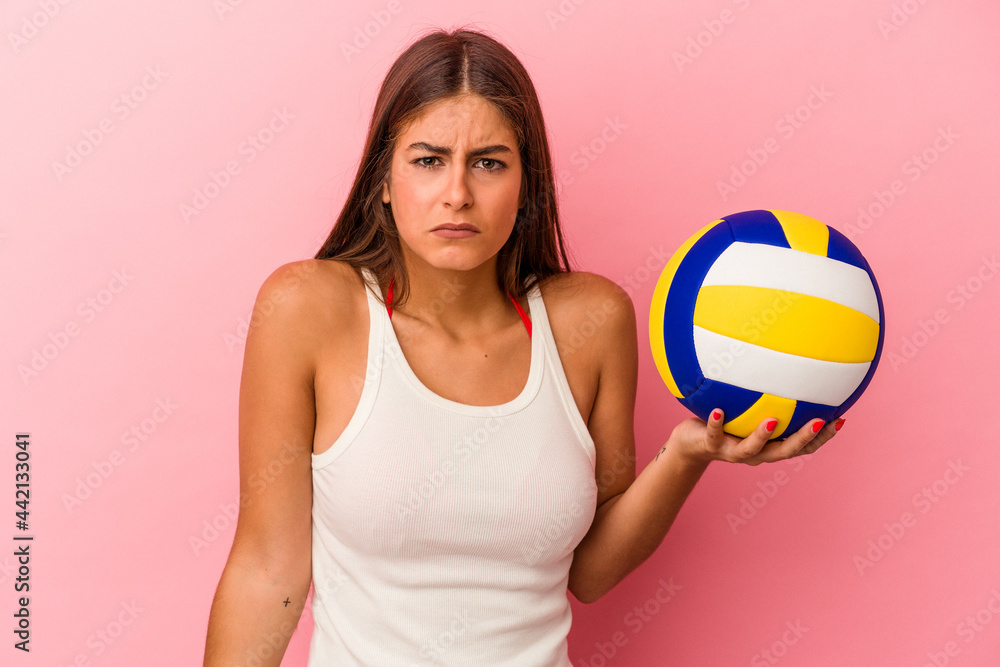 Young caucasian woman holding a volleyball ball isolated on pink background shrugs shoulders and open eyes confused.